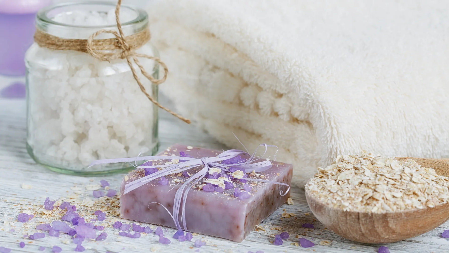 Handmade lavender and oatmeal soap with bath salts, rolled oats, and a soft towel on a wooden surface.