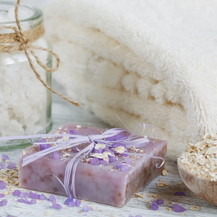 Handmade lavender and oatmeal soap with bath salts, rolled oats, and a soft towel on a wooden surface.