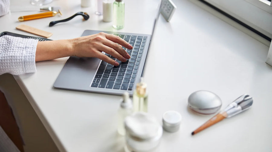 Hand typing on a laptop beside skincare bottles and jars, preparing a MoCRA product listing and facility registration in FDA Cosmetics Direct
